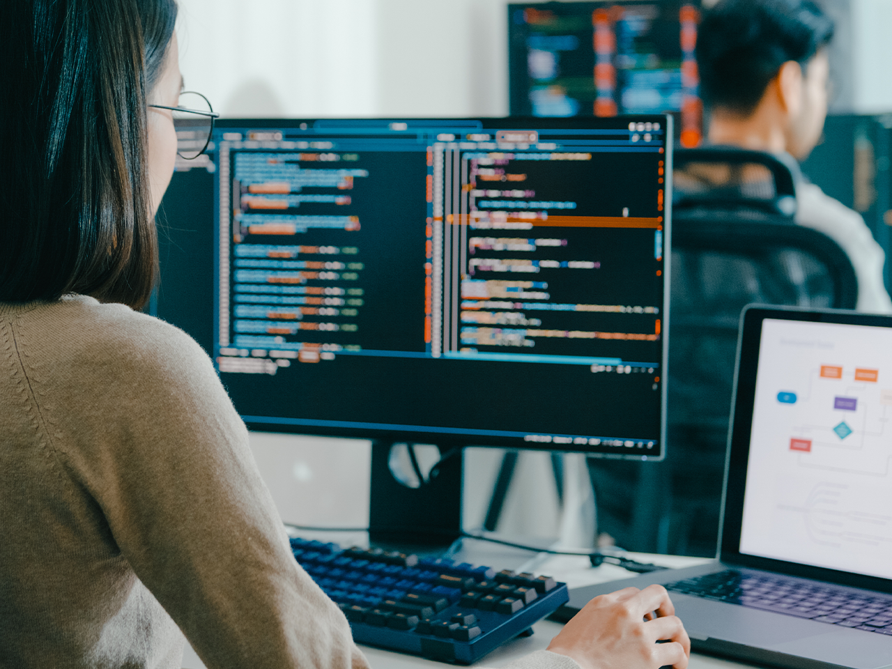 Person wearing glasses sits at a desk with two large monitors showing code; a laptop shows a diagram, and another person is in the background.