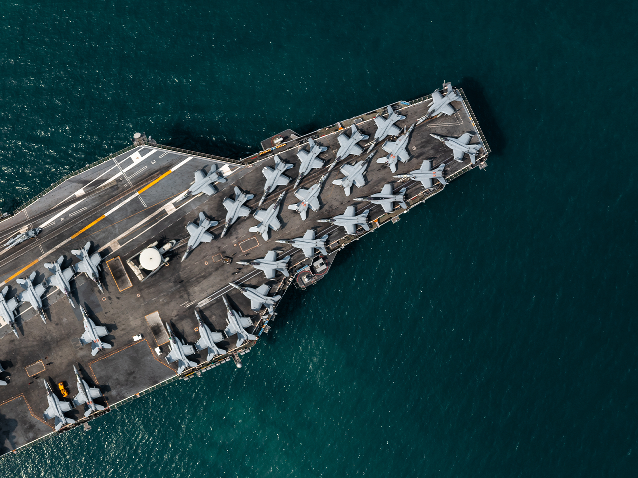 Aerial view of a naval aircraft carrier at sea, its flight deck lined with rows of grey fighter jets, surrounded by deep blue water.