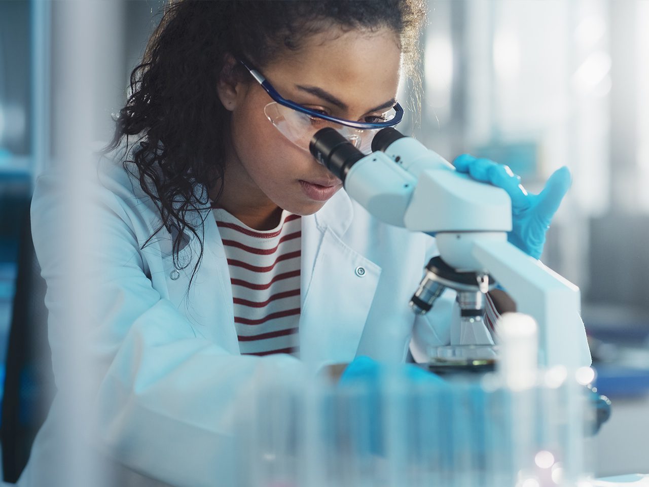 A researcher wearing a lab coat, striped shirt, safety goggles and blue gloves looks through a microscope in a bright laboratory.