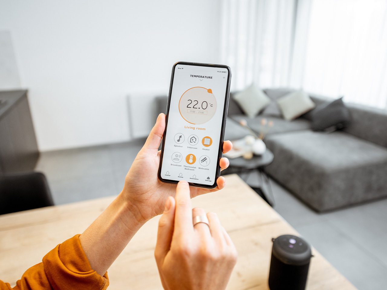 Person holding a smartphone with a control app for a smart home thermostat, showing the temperature set to 22°C in a modern living room.