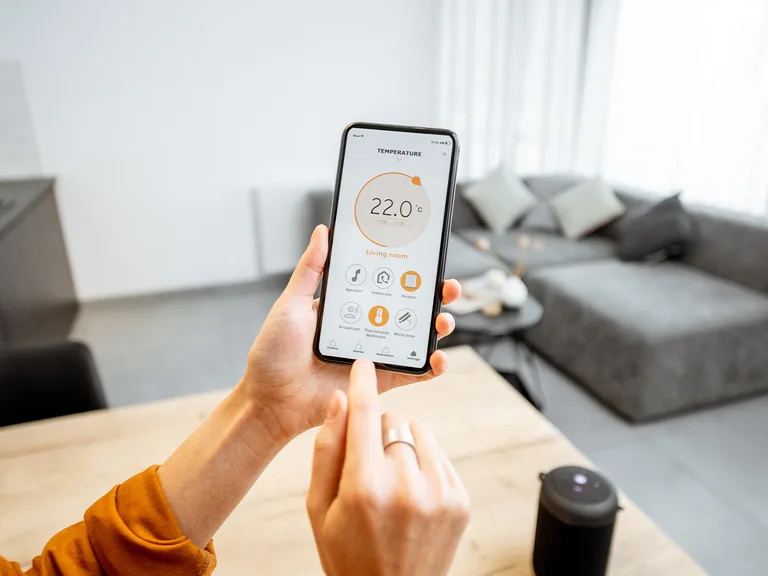 Person holding a smartphone with a control app for a smart home thermostat, showing the temperature set to 22°C in a modern living room.