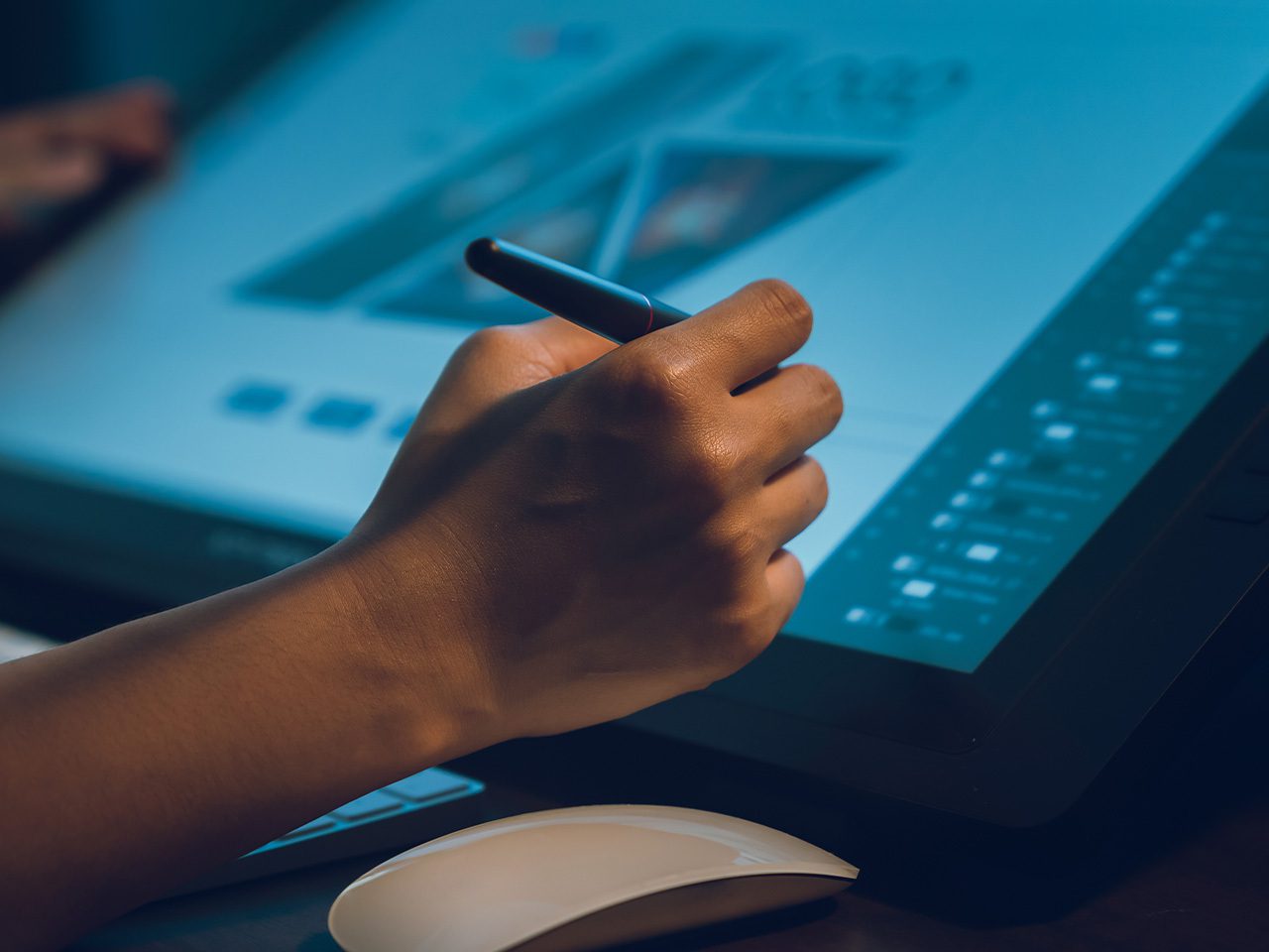 A hand holding a stylus over a digital drawing tablet displaying a design or illustration with a colour palette.