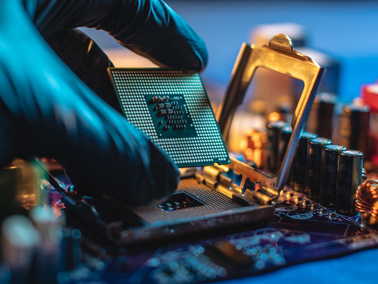 A gloved hand installing a CPU into a computer motherboard, with various electronic components and heating pipes visible in the background.