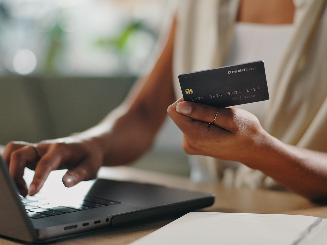 Person typing on a laptop with one hand while holding a black credit card in the other; notebook on the table, blurred background.