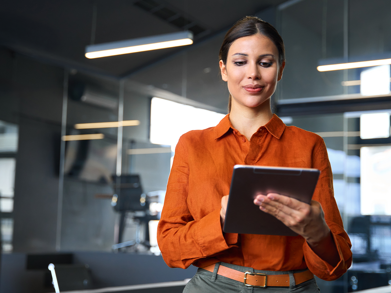 Person in an orange shirt holding a tablet in a modern office, with glass walls and office furniture in the background.