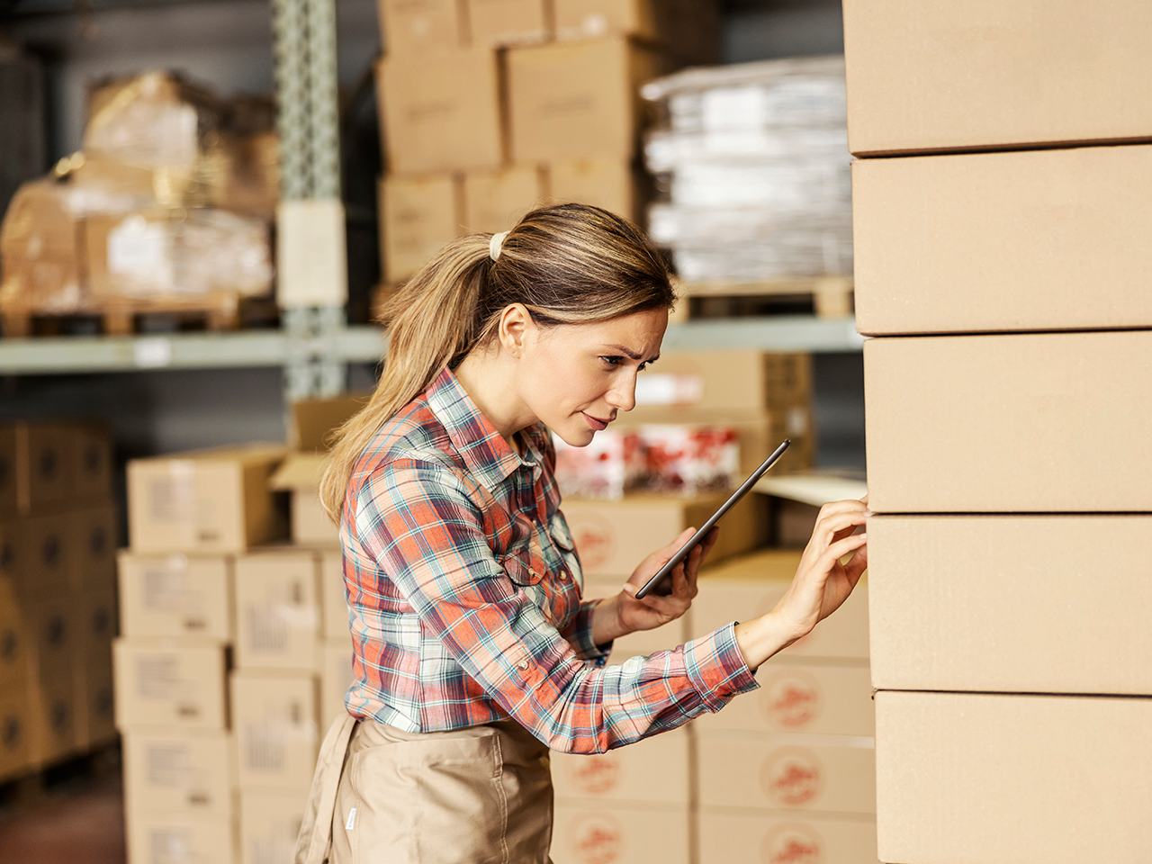 Person in a plaid shirt and beige apron using a tablet, inspecting stacked cardboard boxes in a warehouse.