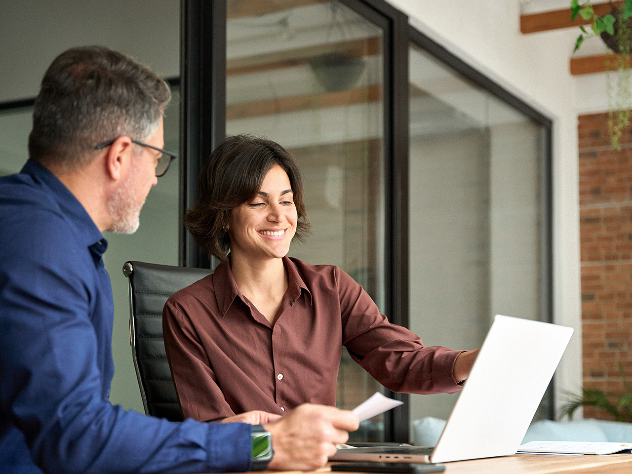 Two people sit at a wooden desk in an office with large windows; one wears a blue shirt and glasses, the other a brown shirt, smiling at a laptop.