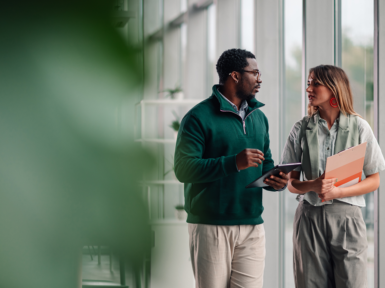 Two professionals stand in a bright glass-walled corridor; one in a dark green fleece with a tablet, the other in a light shirt and pale green vest, holding folders.