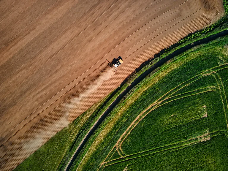 An aerial view of a tractor ploughing a field, leaving a trail of dust, with a lush green area and winding pathways alongside the furrows.