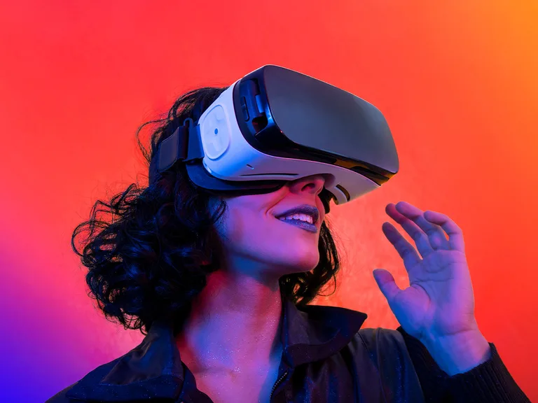 Person wearing a white virtual reality headset with a dark visor, smiling, and raising a hand, against a blue and orange gradient background.
