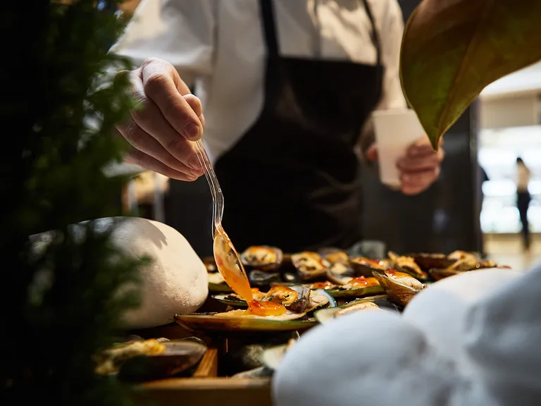 Person in a white shirt and black apron drizzles orange sauce over a platter of mussels and clams; foreground shows a white stone and a large leaf.