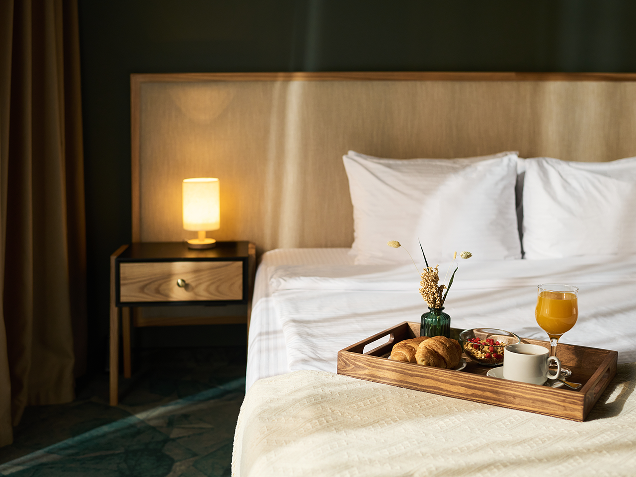 Neatly made bed with white linens and a beige headboard, a bedside lamp, and a breakfast tray on the bed with croissants, juice, coffee and a small vase of dried flowers.