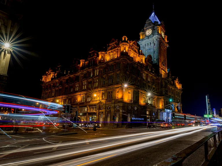 Night view of an ornate clock-tower building bathed in amber light, with long-exposure light trails across the street and a starburst streetlight on the left.