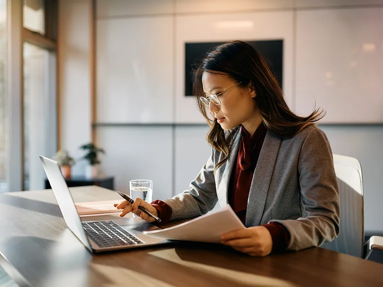 Person with dark hair and glasses sits at a desk in a bright office, working on a laptop, reading papers, with a glass of water nearby.