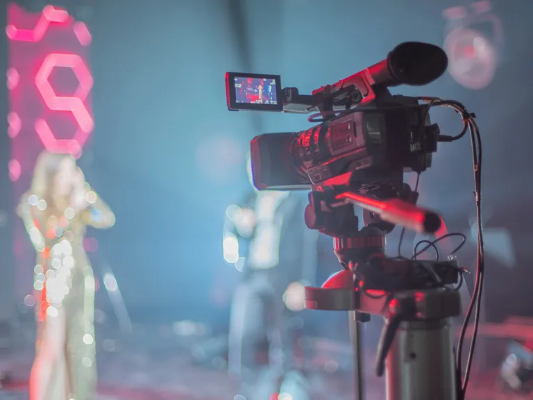 Video camera on a tripod filming a stage performance; a small monitor on the side shows the shot while a glittering performer stands in neon lighting.