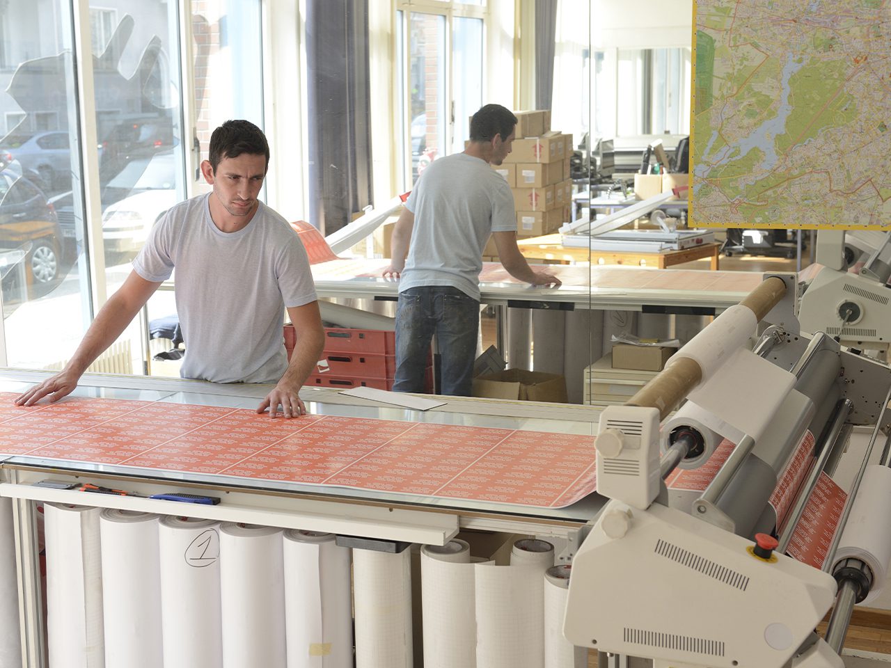 Zwei Personen arbeiten in einer Textilwerkstatt an einer großen Druck-/Schneidemaschine; vorne wird orange bedruckte Stoffbahn ausgelegt, im Hintergrund Kartons und eine Landkarte an der Wand.