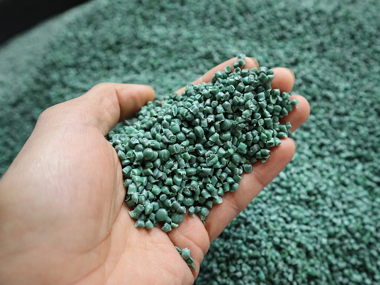 A hand holding small green plastic pellets, with a large pile of similar pellets in the background.