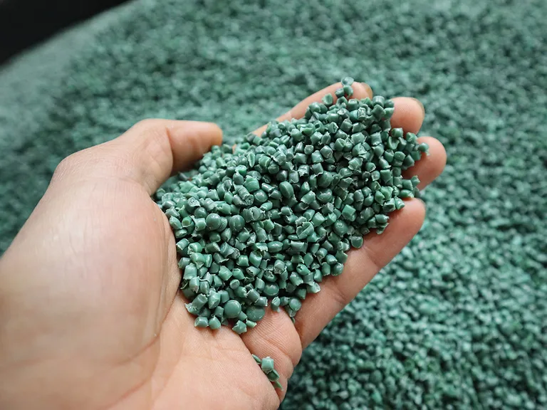 A hand holding small green plastic pellets, with a large pile of similar pellets in the background.