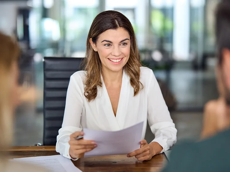 Eine Person in weißer Bluse sitzt an einem Tisch, hält ein Blatt Papier und lächelt während eines Gesprächs in einem modernen Büro.