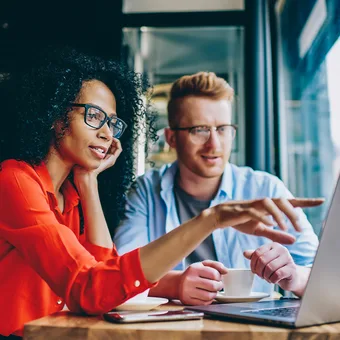 Two people sit at a cafe table with a laptop; one in a red shirt with glasses gestures at the screen while the other, also wearing glasses, listens.