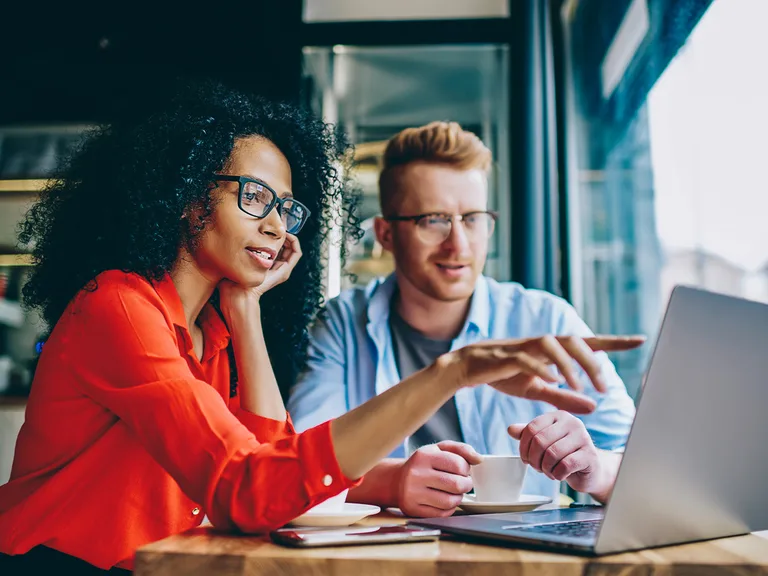 Two people sit at a cafe table with a laptop; one in a red shirt with glasses gestures at the screen while the other, also wearing glasses, listens.