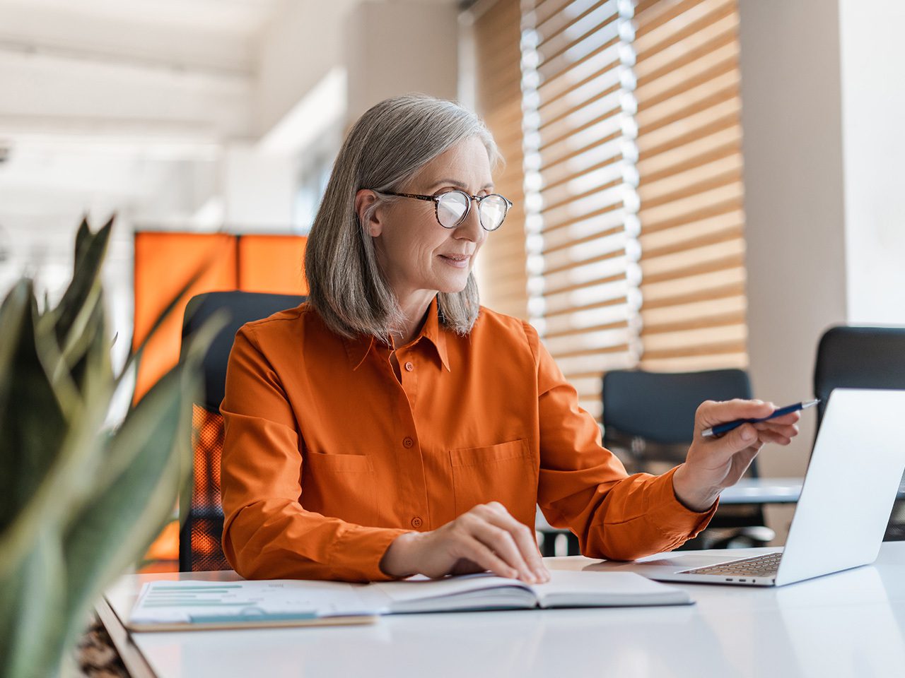 An older person with grey hair and glasses sits at a white desk in a bright office, wearing an orange shirt, with a pen, laptop and open notebook.