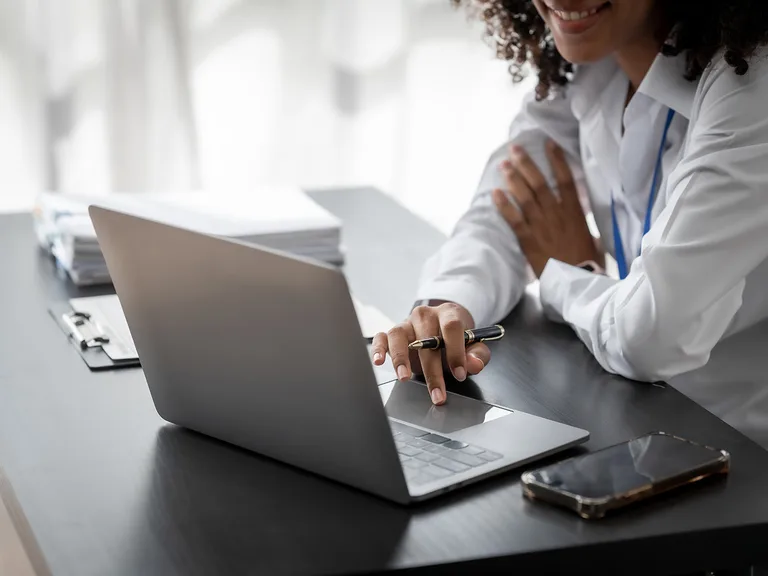 Person in a white shirt sits at a desk with a silver laptop, holding a pen and smiling while working. A smartphone and stack of papers lie nearby.