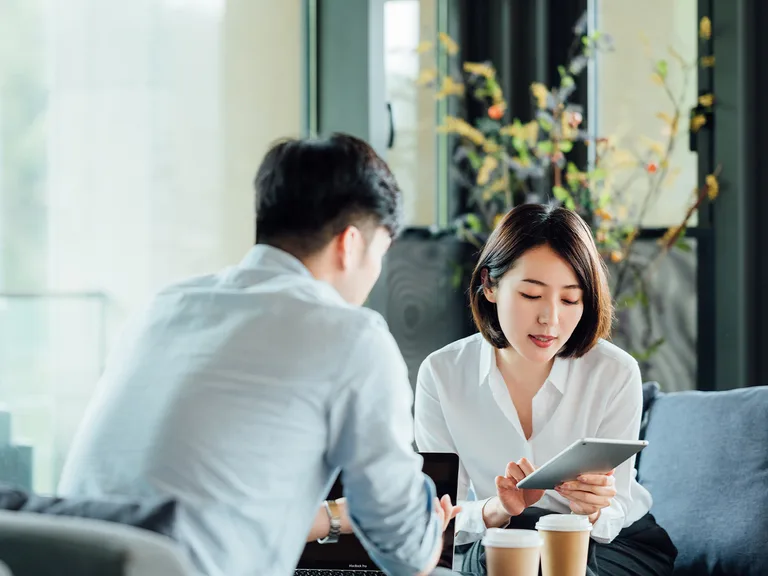 Two professionals in a bright office lounge; one holds a tablet, the other a laptop, with coffee cups on the table and plants in the background.