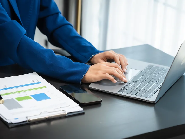 Person in blue blazer working on a laptop, with documents and a smartphone on the desk in a bright office setting.