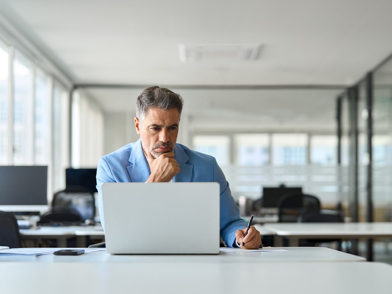 A person in a light blue blazer sits at a desk with a laptop, holding a pen and resting their chin on their hand, in a bright open-plan office.