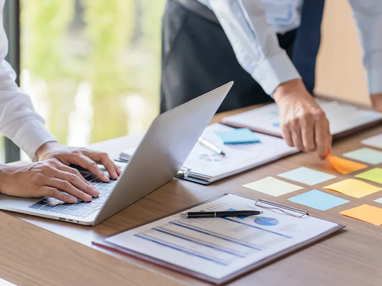 People working at a desk with a laptop, documents, sticky notes, and a pen, engaged in a work or brainstorming session.