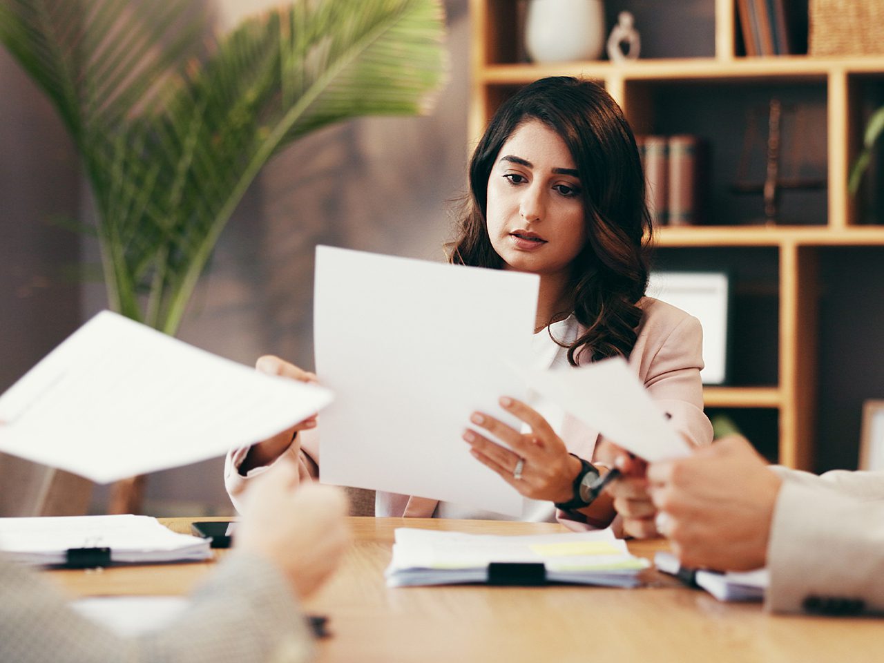 A person with long dark hair sits at a wooden office table, reviewing a document while others pass papers; shelves and a plant behind.