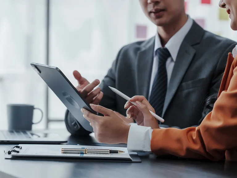 Two colleagues in a business office collaborate over a tablet; one uses a stylus while the other gestures, with a notebook, pen and mug on the table.
