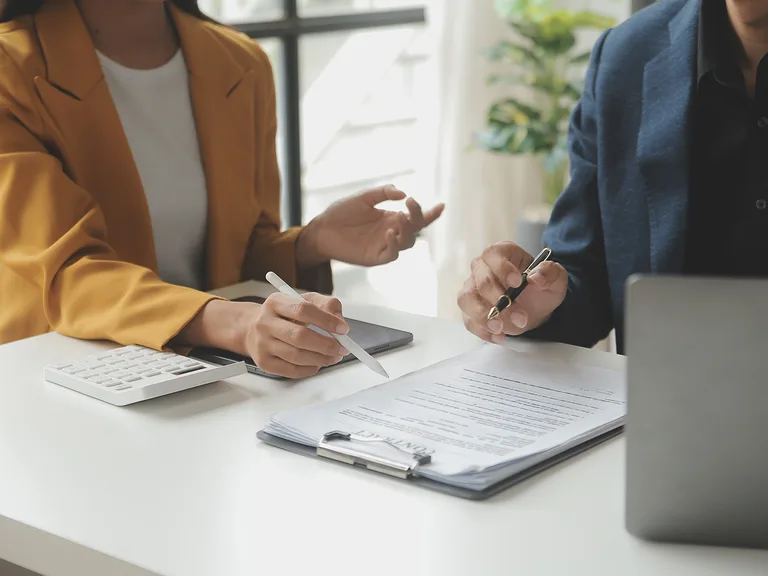 Two people discussing documents at a white table, one with a yellow blazer holding a stylus, the other in a dark blue blazer with a pen, in a bright office setting.
