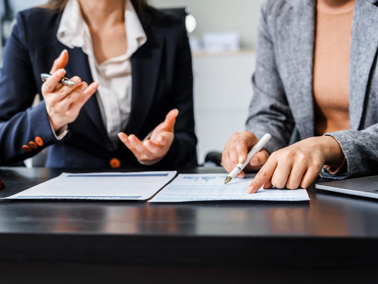 Two professionals in business attire review documents at a desk; one gestures with a pen, the other points at a form, with a laptop nearby.
