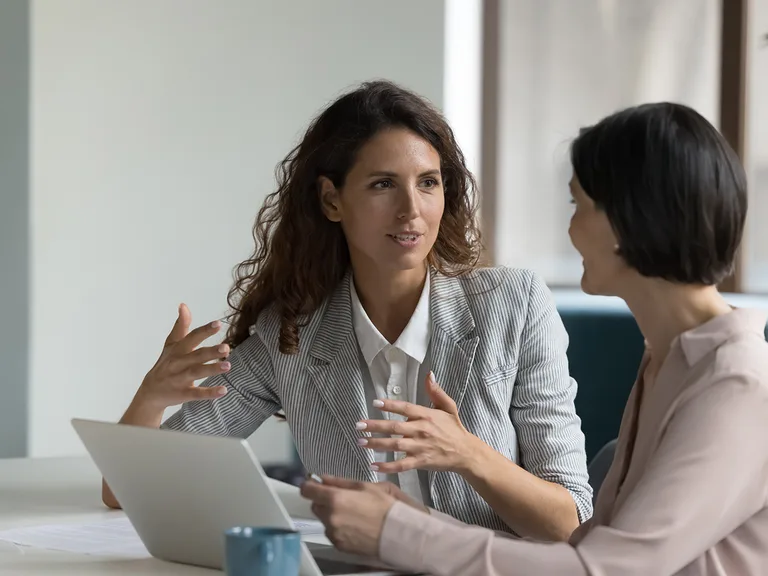 Two women in a bright office discuss at a table with a laptop; one with curly hair in a striped blazer gestures, the other with short dark hair listens.