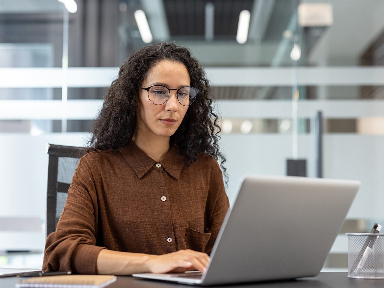 Person with curly dark hair and glasses wearing a brown shirt, seated at a desk and typing on a silver laptop in a glass-partitioned office.