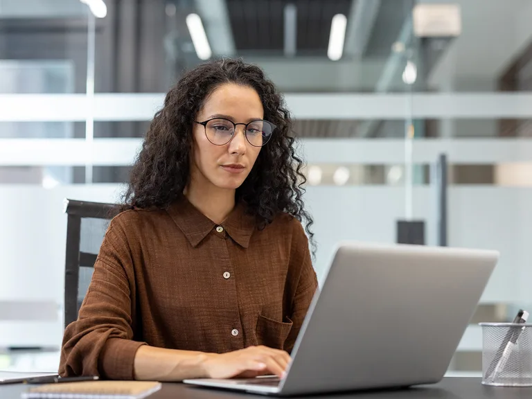 Person with curly dark hair and glasses wearing a brown shirt, seated at a desk and typing on a silver laptop in a glass-partitioned office.