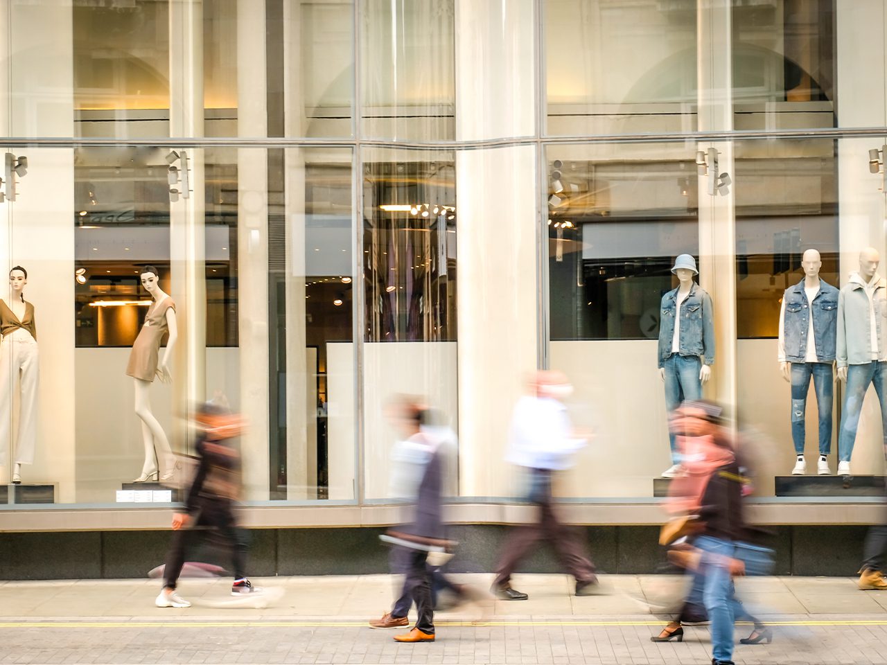 Storefront window display with mannequins in beige and denim outfits behind glass, while blurred pedestrians walk along a city street.
