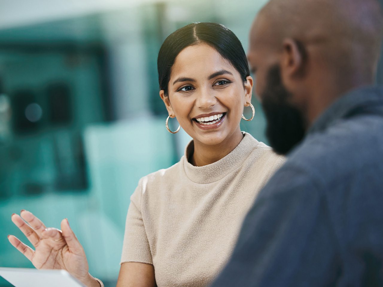 Two people chat in a bright, modern space; a person with dark hair in a beige top and hoop earrings smiles and gestures, while the other has a beard.