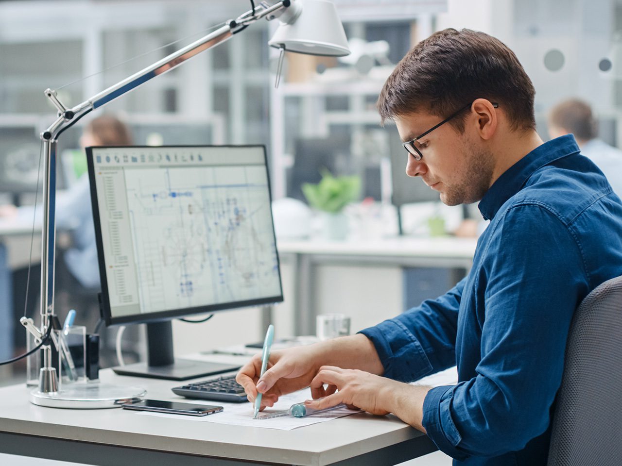 A person in a blue shirt and glasses sits at a desk, writing on papers with a pen as a monitor shows diagrams in a bright office.