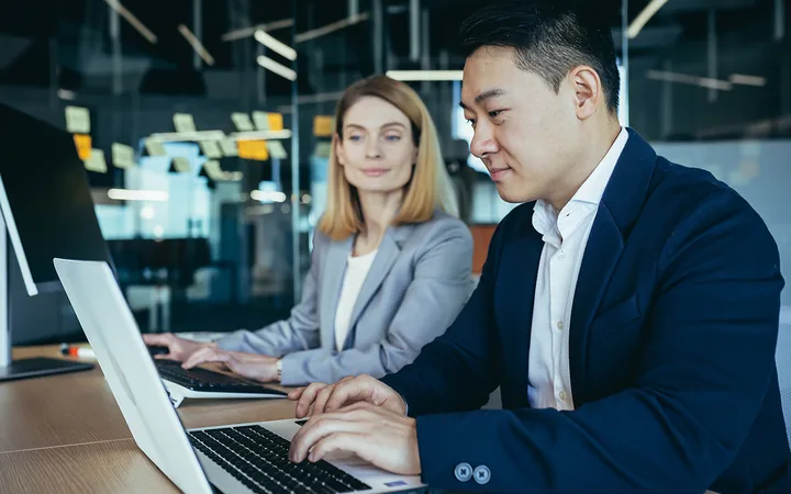 Two professionals sit at a wooden table working on laptops in a modern office; glass walls with sticky notes in the background.