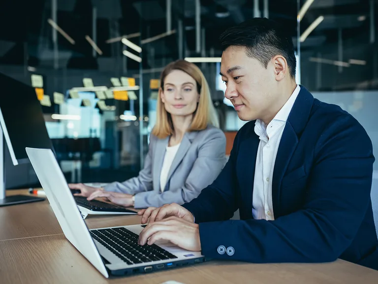 Two professionals sit at a wooden table in a modern office, each typing on a laptop, with a glass wall and sticky notes visible behind.
