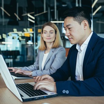 Two professionals sit at a wooden table with laptops in a modern office, with a glass wall behind them covered in sticky notes.