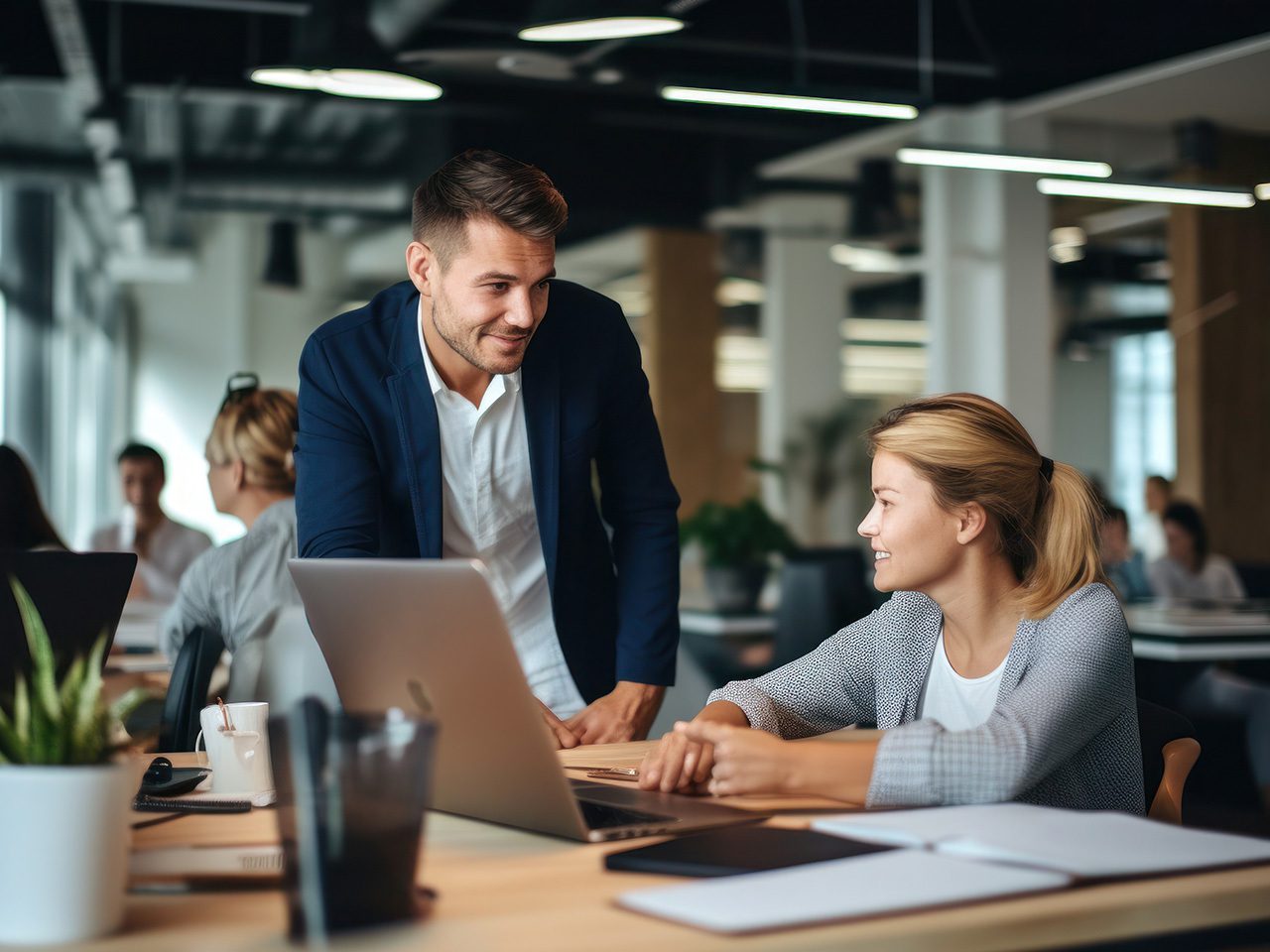 Person standing and leaning over a desk to discuss a laptop with a seated colleague in a bright, modern open-plan office, with other staff in the background.