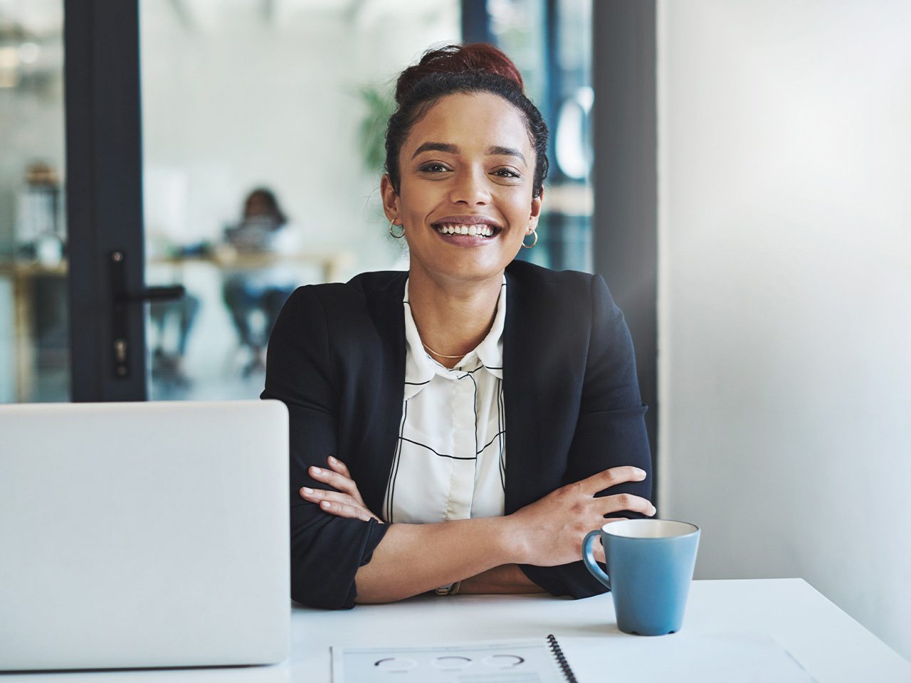 Una persona sorridente, in giacca nera e camicia bianca, seduta al tavolo con laptop aperto e una tazza blu in un ufficio moderno.