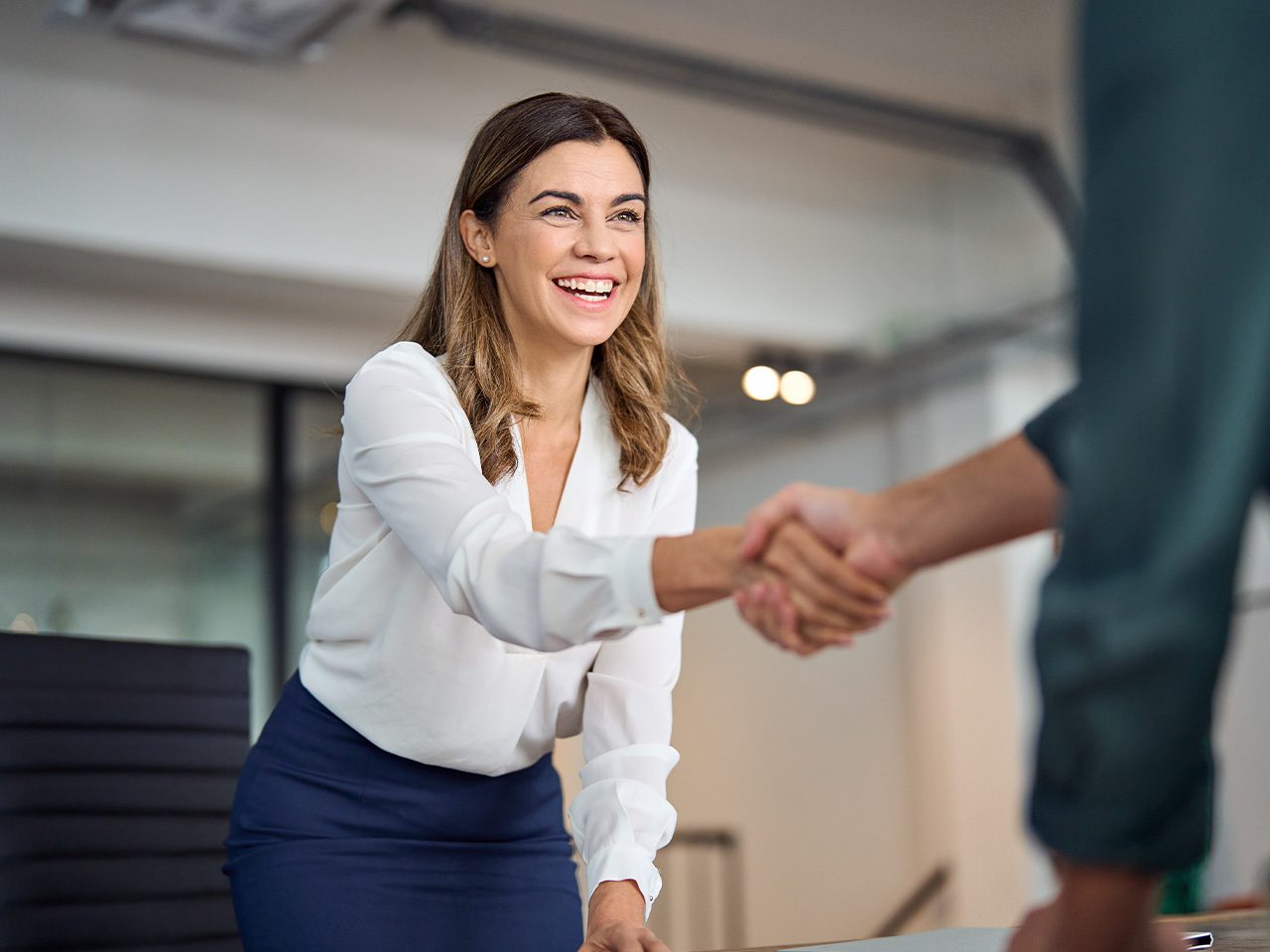 A smiling person in a white blouse and navy skirt shakes hands with a colleague in a bright office setting.