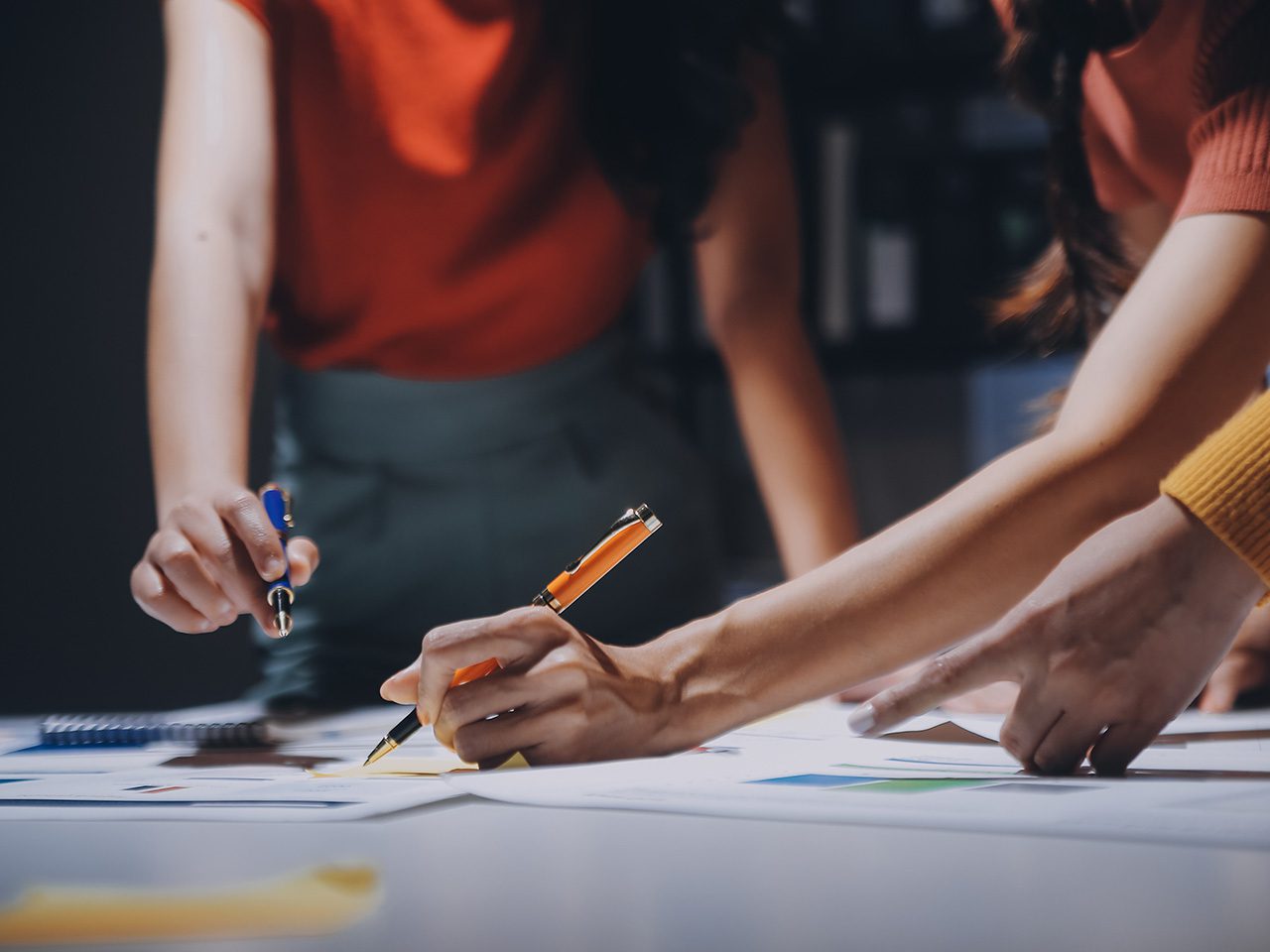 Close-up of several hands writing on papers with blue and orange pens at a collaborative workspace.