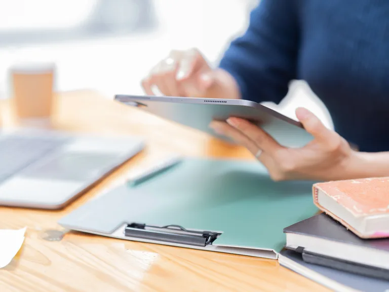 Person using a tablet at a desk surrounded by notebooks, documents, and a calculator, with a coffee cup in the background.