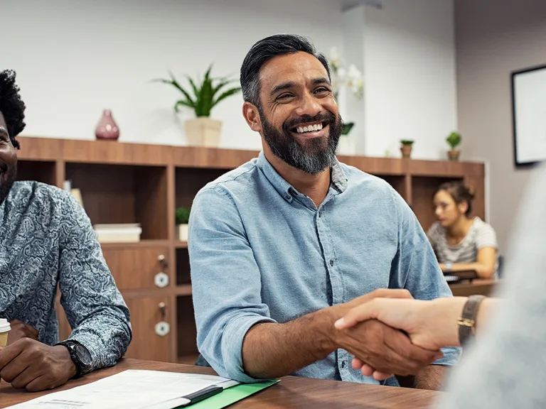 A bearded person in a light blue shirt smiles while shaking hands across a table in a bright office; another colleague sits nearby.